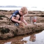 Een jonge jongen speelt met een schep en een emmer op het strand bij water, genietend van buitenactiviteiten.