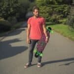 Skateboard met kleurrijk design en groene wielen, man staat op weg met skateboard in hand.