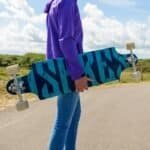 Kleurrijke longboard met graffiti-ontwerp, vastgehouden door een persoon in een paarse jas. Buitenland landschap achter, perfect voor skaten of cruisen.