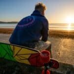 Een man zit aan het strand tijdens zonsondergang met zijn skateboard, rustgevend en geschikt voor outdoor skateboardplezier.