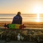 Skateboarder zit aan zee tijdens zonsondergang met skateboard op de rand van een muur.