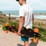 Korte skateboard met oranje wielen in de hand van een man bij de kust.