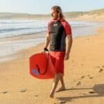 Strandwandelaar met surfboard op het strand met branding en duinen op de achtergrond.
