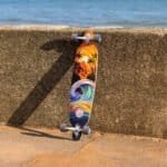 Kleurrijk skateboard met tropisch zonsondergang design, leunend tegen betonnen muur aan strand.