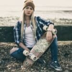 Skateboard vrouw op strand met hoodie, ripped jeans en beanie, klaar voor buitensportplezier.